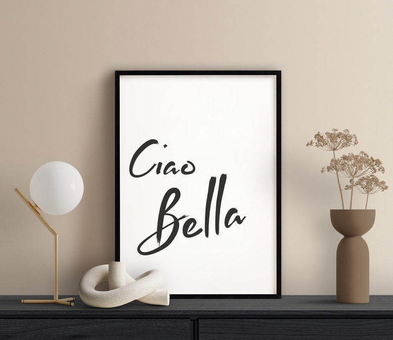 May include: Black and white framed print with the text "Ciao Bella" in a handwritten style. The print is displayed on a black shelf with a white ceramic sculpture, a gold lamp, and a brown vase with dried flowers.