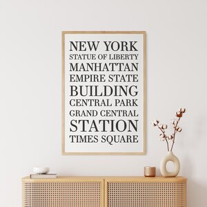 May include: A framed print with black text on a white background. The text reads "NEW YORK", "STATUE OF LIBERTY", "MANHATTAN", "EMPIRE STATE BUILDING", "CENTRAL PARK", "GRAND CENTRAL STATION", and "TIMES SQUARE".
