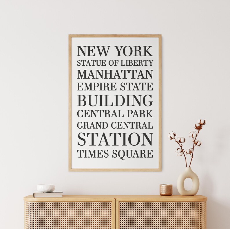 May include: A framed print with black text on a white background. The text reads "NEW YORK", "STATUE OF LIBERTY", "MANHATTAN", "EMPIRE STATE BUILDING", "CENTRAL PARK", "GRAND CENTRAL STATION", and "TIMES SQUARE".