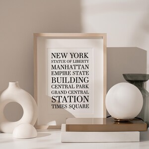 May include: A framed print with the text "NEW YORK" and a list of iconic New York City landmarks, including the Statue of Liberty, Manhattan, Empire State Building, Central Park, Grand Central Station, and Times Square. The print is displayed on a white surface with a white ceramic vase and a white globe lamp.
