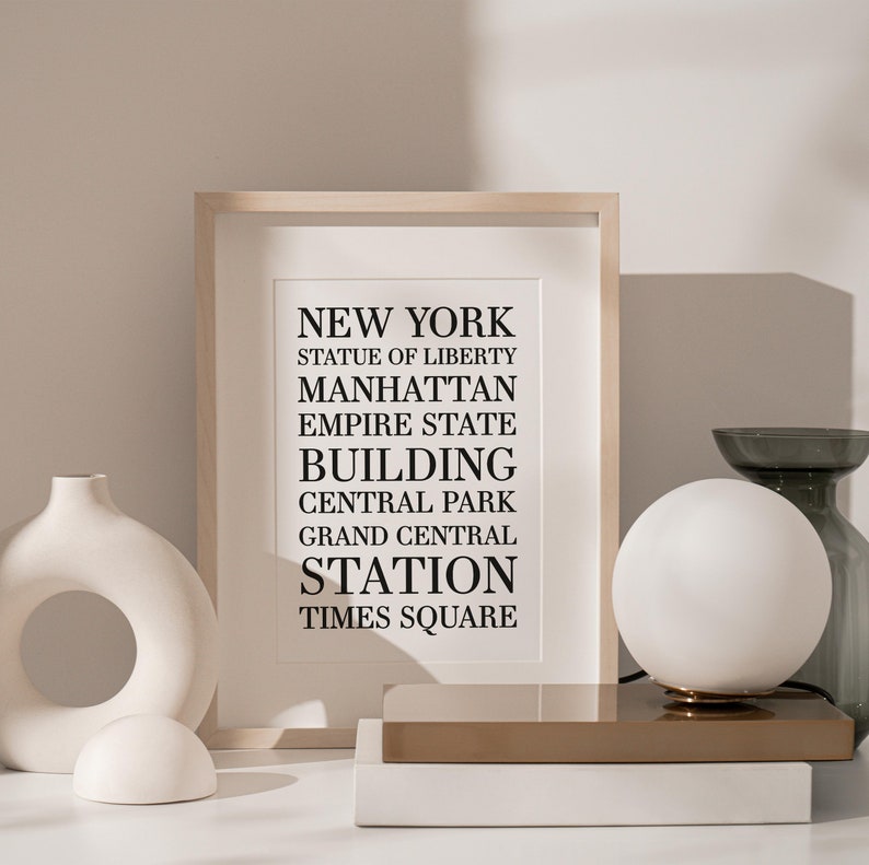 May include: A framed print with the text "NEW YORK" and a list of iconic New York City landmarks, including the Statue of Liberty, Manhattan, Empire State Building, Central Park, Grand Central Station, and Times Square. The print is displayed on a white surface with a white ceramic vase and a white globe lamp.