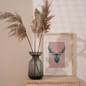 May include: A gray glass vase with dried pampas grass and a framed print of a geometric deer on a light pink background. The vase and print are on a wooden shelf with a woven rattan front.