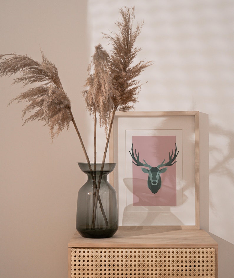 May include: A gray glass vase with dried pampas grass and a framed print of a geometric deer on a light pink background. The vase and print are on a wooden shelf with a woven rattan front.