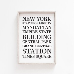 May include: A framed print with the text "NEW YORK" in black lettering, followed by a list of iconic New York City landmarks: Statue of Liberty, Manhattan, Empire State Building, Central Park, Grand Central Station, and Times Square.