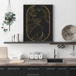 May include: A black and gold framed print of a line drawing of a woman's face. The woman is looking to the right and has her hand on her chin. The background is black and the lines are gold.