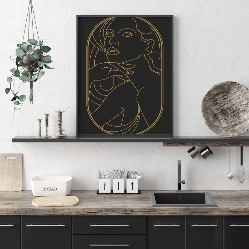 May include: A black and gold framed print of a line drawing of a woman's face. The woman is looking to the right and has her hand on her chin. The background is black and the lines are gold.