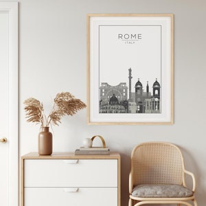 May include: A black and white illustration of the Rome skyline with the text "ROME - ITALY" above the illustration. The illustration is framed in a light wood frame and is hanging on a white wall. The wall is next to a white dresser with three drawers and a light wood top. A vase with dried pampas grass is on the dresser. A gold metal arch is on the dresser next to the vase. A wicker chair with a gray cushion is in the foreground.