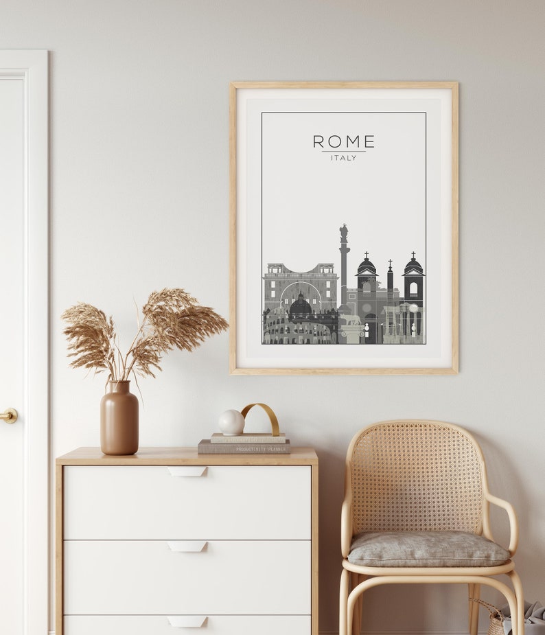 May include: A black and white illustration of the Rome skyline with the text "ROME - ITALY" above the illustration. The illustration is framed in a light wood frame and is hanging on a white wall. The wall is next to a white dresser with three drawers and a light wood top. A vase with dried pampas grass is on the dresser. A gold metal arch is on the dresser next to the vase. A wicker chair with a gray cushion is in the foreground.