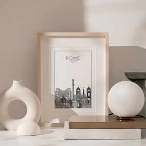 May include: A framed print of a city skyline with the text "ROME ITALY" above the image. The print is on a white background and is framed in a light wood frame. The print is sitting on a white surface with a white ceramic vase and a white globe lamp.