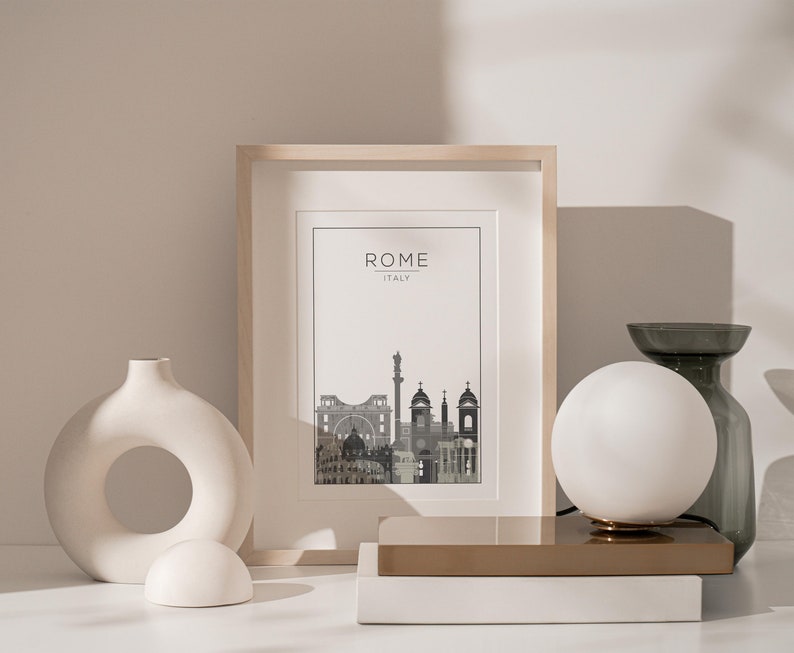 May include: A framed print of a city skyline with the text "ROME ITALY" above the image. The print is on a white background and is framed in a light wood frame. The print is sitting on a white surface with a white ceramic vase and a white globe lamp.