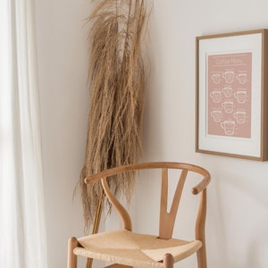 May include: A framed print with a pink background and white text that reads "Coffee Menu" with illustrations of coffee cups. The print is hanging on a white wall next to a wooden chair with a woven seat.