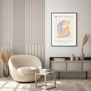 May include: A beige armchair with a modern design sits in a room with a white wall and a wooden floor. A black framed poster with the text "MATISSE GALERIE D'EXPOSITION Papier Decoup&eacute;" hangs on the wall.