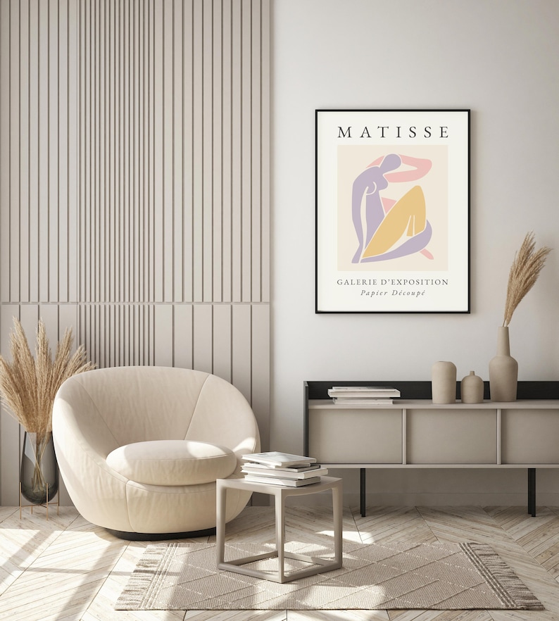May include: A beige armchair with a modern design sits in a room with a white wall and a wooden floor. A black framed poster with the text "MATISSE GALERIE D'EXPOSITION Papier Decoup&eacute;" hangs on the wall.