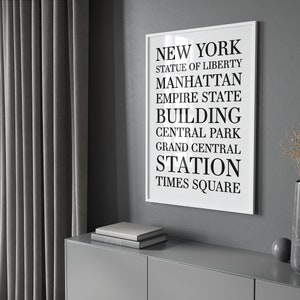 May include: A black and white typography print with the words "NEW YORK", "STATUE OF LIBERTY", "MANHATTAN", "EMPIRE STATE BUILDING", "CENTRAL PARK", "GRAND CENTRAL STATION", and "TIMES SQUARE" in a white frame against a gray wall.