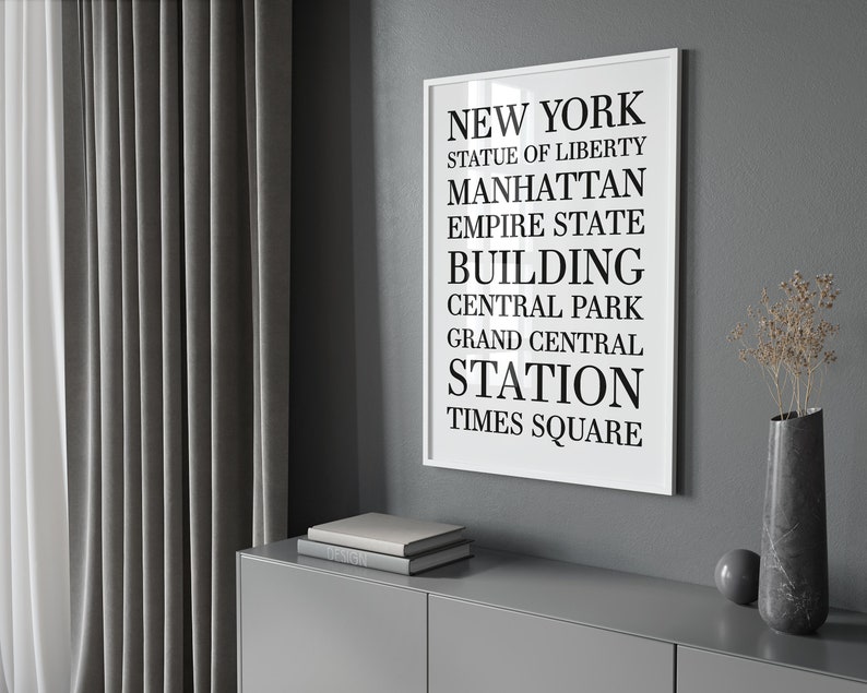 May include: A black and white typography print with the words "NEW YORK", "STATUE OF LIBERTY", "MANHATTAN", "EMPIRE STATE BUILDING", "CENTRAL PARK", "GRAND CENTRAL STATION", and "TIMES SQUARE" in a white frame against a gray wall.