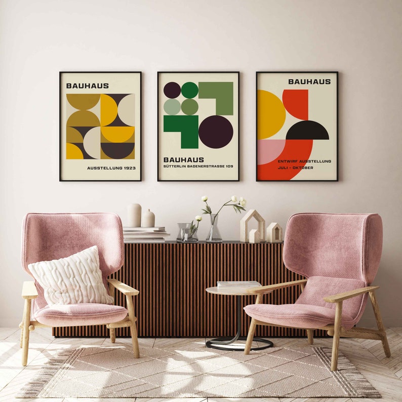 May include: Three framed Bauhaus posters with geometric designs in yellow, red, green, and black on a cream background. The posters are hanging on a white wall above a wooden cabinet and two pink armchairs.