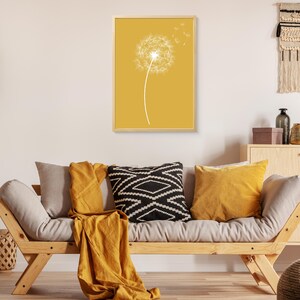 Op de afbeelding: Een ingelijste print van een witte paardenbloem met zaden die in de wind waaien op een mosterdgele achtergrond.
