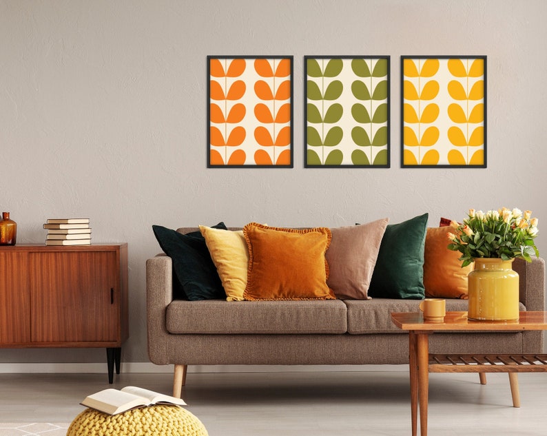 May include: Three framed prints with a retro pattern of orange, yellow, and green leaves on a white background. The prints are hanging on a light gray wall above a brown sofa with pillows in shades of green, brown, and orange. A wooden coffee table sits in front of the sofa.
