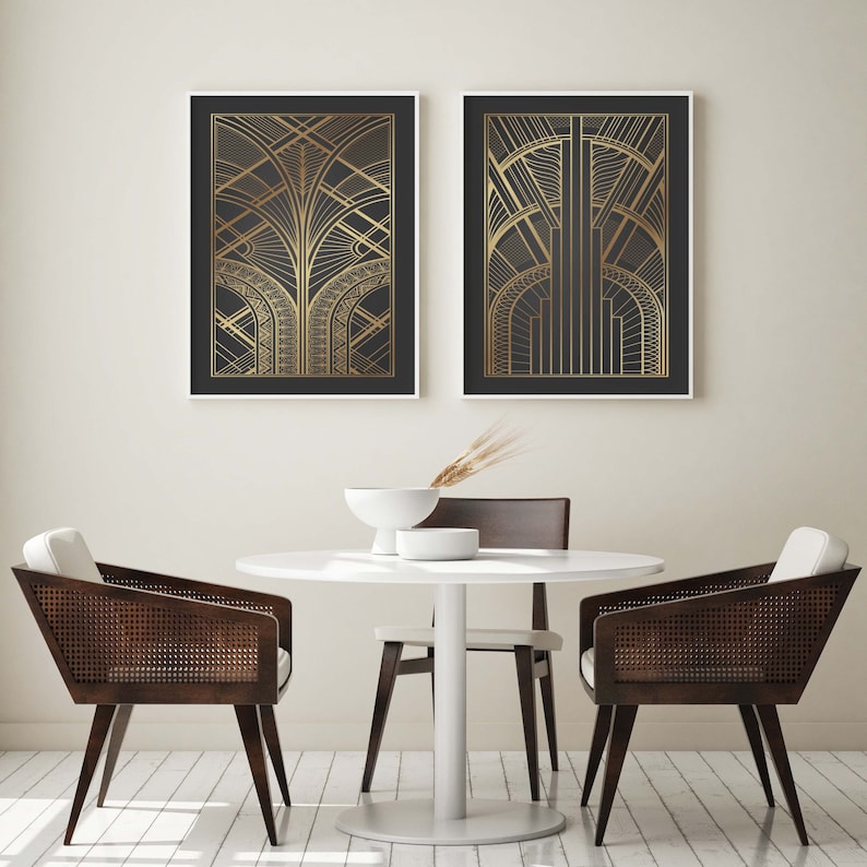 May include: Two black framed prints with gold geometric designs hang on a white wall above a white round table with four brown chairs. The table is set with a white bowl and a vase with dried grass.