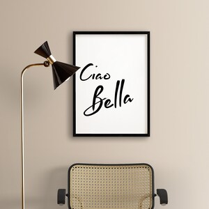 May include: Black framed print with white background and the text "Ciao Bella" in black script font. The print is hanging on a beige wall above a wicker chair.