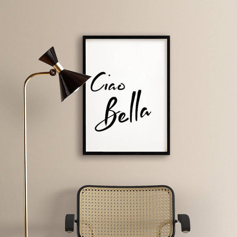 May include: Black framed print with white background and the text "Ciao Bella" in black script font. The print is hanging on a beige wall above a wicker chair.