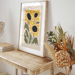 May include: A framed print of a stylized sunflower design with yellow petals and black centers. The print is on a beige background and is framed in a light wood frame.