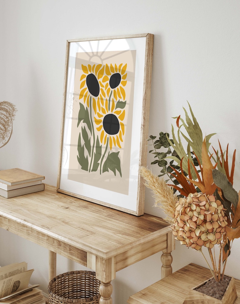 May include: A framed print of a stylized sunflower design with yellow petals and black centers. The print is on a beige background and is framed in a light wood frame.