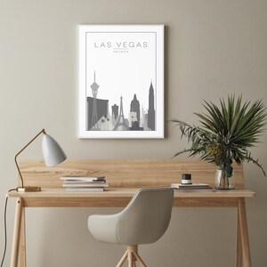 K&ouml;nnte beinhalten: Ein gerahmter Druck einer grauen Skyline mit dem Text "LAS VEGAS, NEVADA" oben. Der Druck h&auml;ngt an einer hellgrauen Wand &uuml;ber einem Holztisch mit einer Lampe, einem Stuhl und einer Vase mit Gr&uuml;npflanzen.