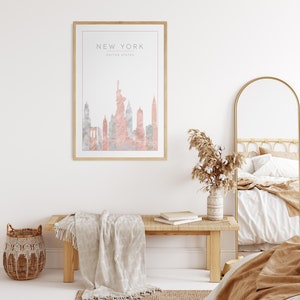 May include: A framed print with a watercolor illustration of the New York City skyline in pink and gray tones. The print is hanging on a white wall above a wooden bench with a white throw blanket.