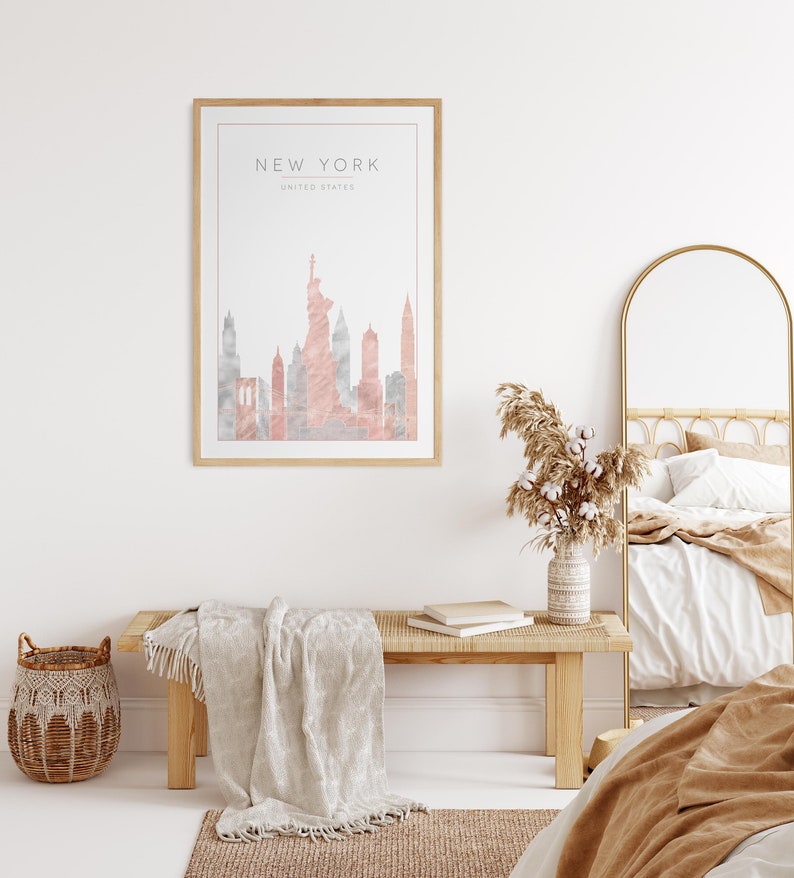May include: A framed print with a watercolor illustration of the New York City skyline in pink and gray tones. The print is hanging on a white wall above a wooden bench with a white throw blanket.