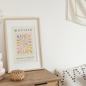 May include: A framed print of a Matisse painting titled "Musee d'Orsay" featuring a colorful abstract design of pink, yellow, and purple shapes. The print is in a light wood frame and is hanging on a white wall.