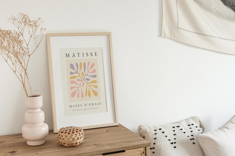 May include: A framed print of a Matisse painting titled "Musee d'Orsay" featuring a colorful abstract design of pink, yellow, and purple shapes. The print is in a light wood frame and is hanging on a white wall.