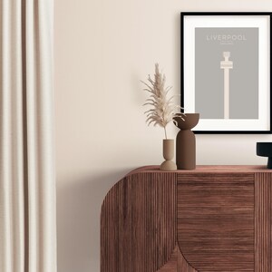 May include: A framed print with a minimalist design of a tall building with the text "LIVERPOOL ENGLAND" above it. The print is on a gray background and is framed in black. The print is sitting on a wooden cabinet with two small vases and a dried plant.