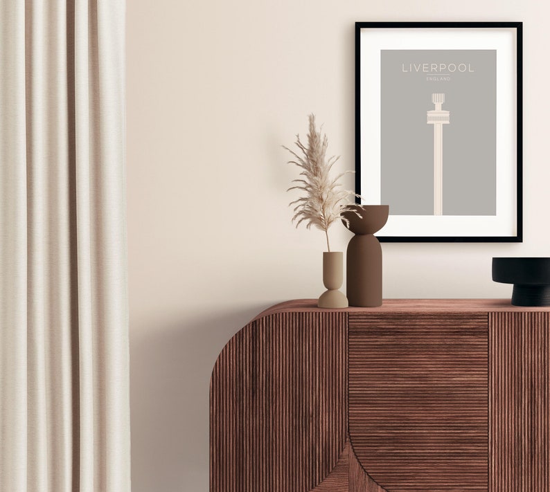 May include: A framed print with a minimalist design of a tall building with the text "LIVERPOOL ENGLAND" above it. The print is on a gray background and is framed in black. The print is sitting on a wooden cabinet with two small vases and a dried plant.