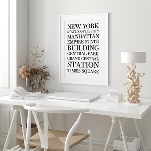May include: A white framed print with black text listing iconic New York City landmarks and locations, including the Statue of Liberty, Manhattan, Empire State Building, Central Park, Grand Central Station, and Times Square. The print is displayed on a white wall above a white desk with a laptop, a vase of dried flowers, and a lamp.
