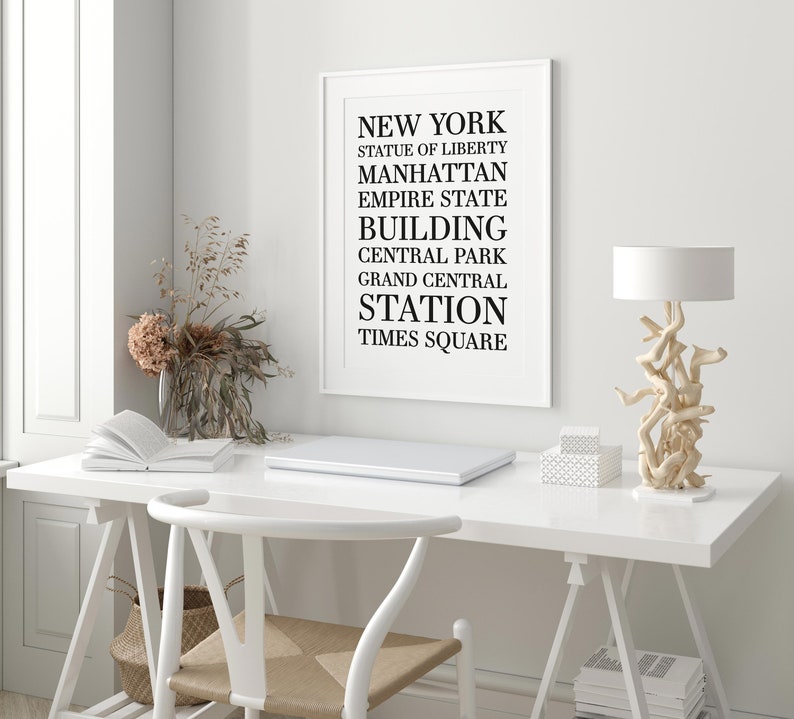 May include: A white framed print with black text listing iconic New York City landmarks and locations, including the Statue of Liberty, Manhattan, Empire State Building, Central Park, Grand Central Station, and Times Square. The print is displayed on a white wall above a white desk with a laptop, a vase of dried flowers, and a lamp.