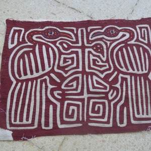 May include: A maroon fabric wall hanging with a white embroidered design of two stylized birds with their beaks touching. The birds are surrounded by a geometric pattern of white lines.