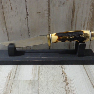 May include: A hunting knife with a brown and white stag handle and a black wooden display stand.