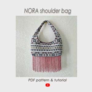 Peut inclure: NORA shoulder bag PDF pattern & tutorial. Un sac à bandoulière en forme de croissant avec un motif géométrique inspiré des flocons de neige en blanc, noir et rouge, agrémenté d'une longue frange rose unie sur le bord inférieur.