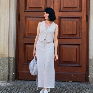 Puede incluir: Una mujer lleva un conjunto de chaleco y falda de lino a rayas blancas y marrones. Est&aacute; de pie frente a una puerta de madera y lleva un bolso gris.