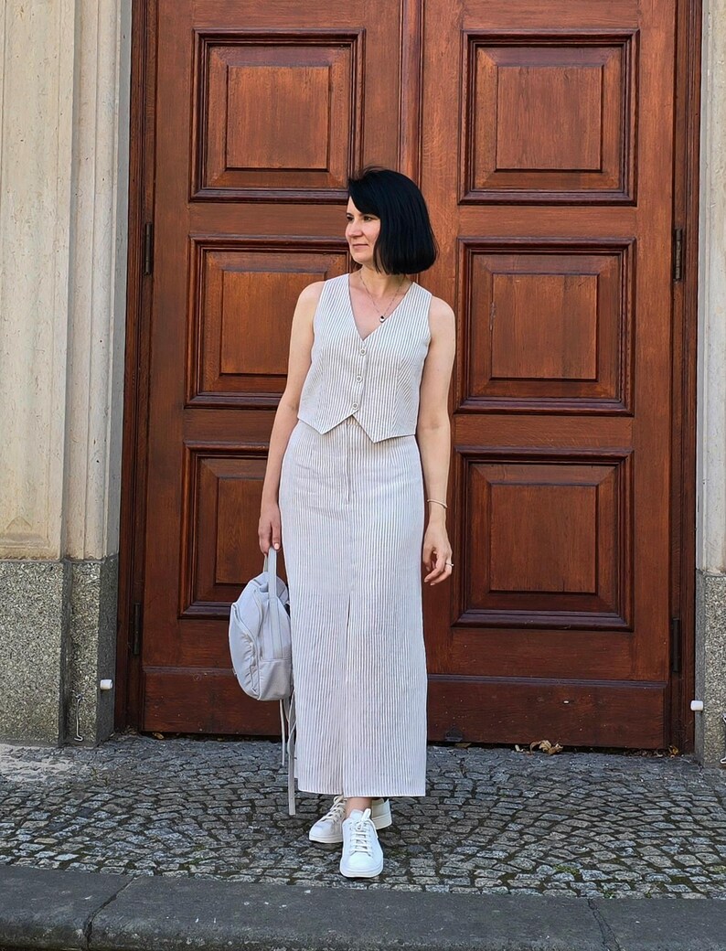 Puede incluir: Una mujer lleva un conjunto de chaleco y falda de lino a rayas blancas y marrones. Est&aacute; de pie frente a una puerta de madera y lleva un bolso gris.