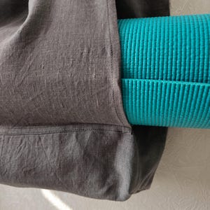 May include: A gray linen tote bag with a teal yoga mat tucked inside. The bag has a pocket on the side and a stitched seam across the bottom.