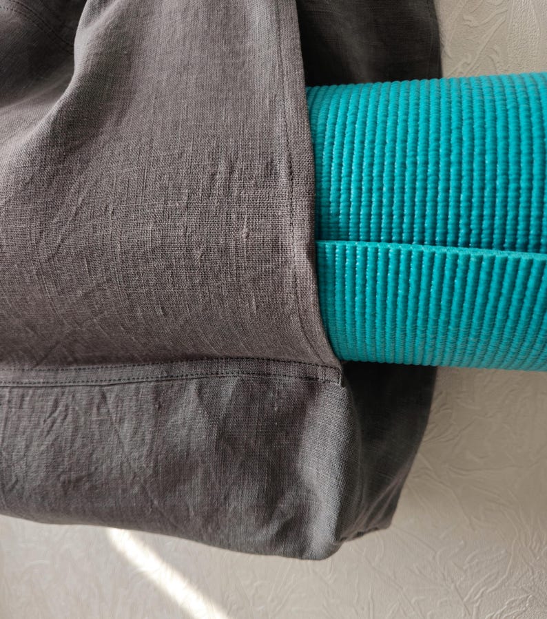 May include: A gray linen tote bag with a teal yoga mat tucked inside. The bag has a pocket on the side and a stitched seam across the bottom.