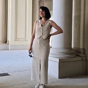 Puede incluir: Una mujer lleva un chaleco sin mangas beige y una falda a juego con una abertura. Est&aacute; de pie frente a una columna blanca y lleva zapatillas blancas.
