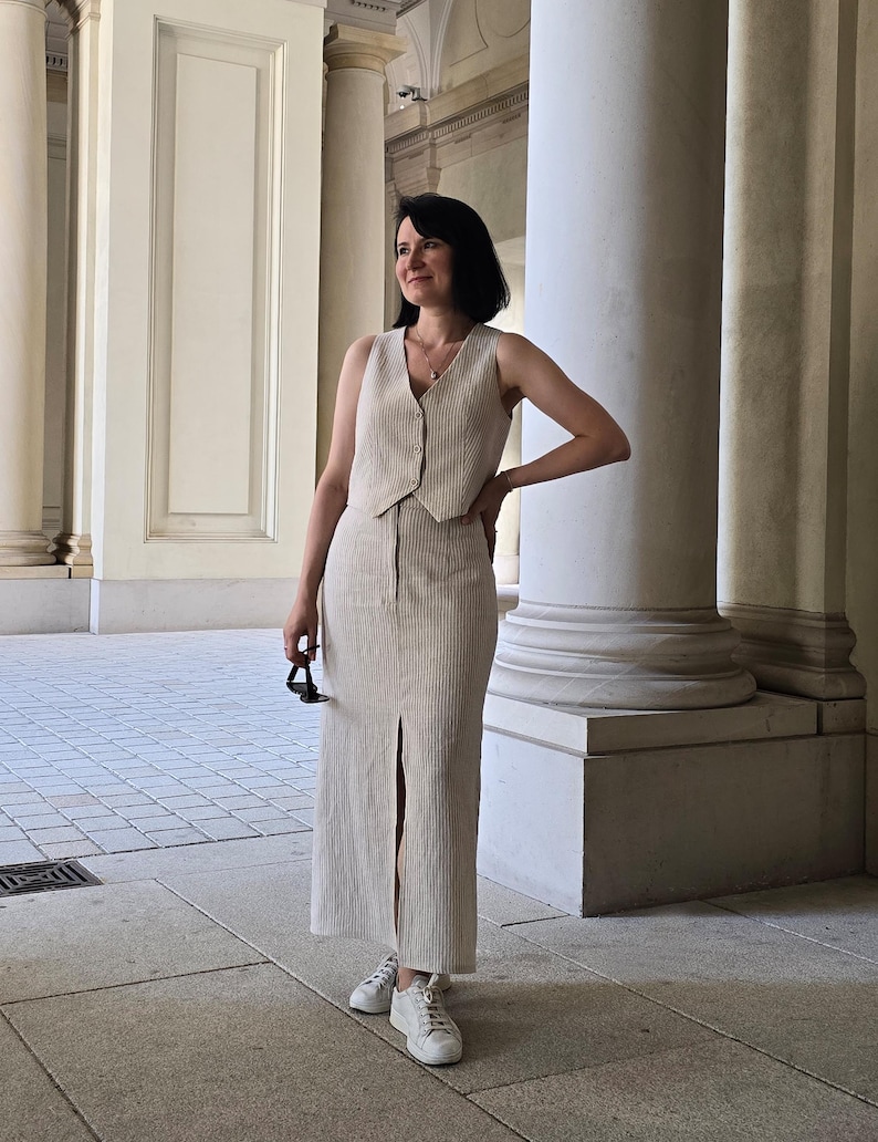Puede incluir: Una mujer lleva un chaleco sin mangas beige y una falda a juego con una abertura. Est&aacute; de pie frente a una columna blanca y lleva zapatillas blancas.