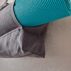 May include: A gray linen tote bag with a teal yoga mat tucked inside. The bag has a single strap and a zipper closure.