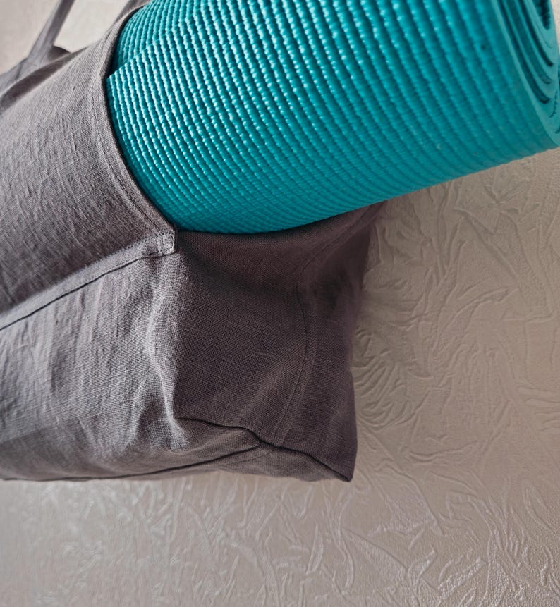 May include: A gray linen tote bag with a teal yoga mat tucked inside. The bag has a single strap and a zipper closure.