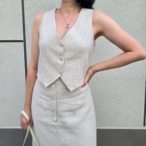 Puede incluir: Un conjunto de chaleco y falda de lino a rayas en color crema y beige. El chaleco tiene cuello en V y botones en la parte delantera. La falda es de largo midi y tiene una silueta recta.