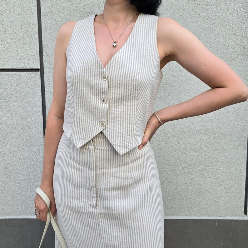 Puede incluir: Un conjunto de chaleco y falda de lino a rayas en color crema y beige. El chaleco tiene cuello en V y botones en la parte delantera. La falda es de largo midi y tiene una silueta recta.