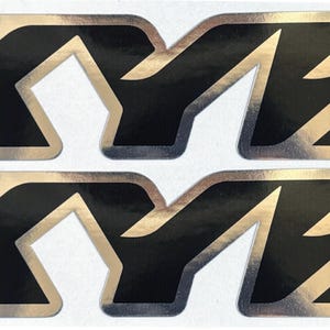 Puede incluir: Dos pegatinas plateadas y negras con el logotipo KYB. Las pegatinas presentan las letras "KYB" en una fuente audaz y estilizada. Las letras son negras con un contorno plateado. Para aplicaciones automotrices o motocicletas.